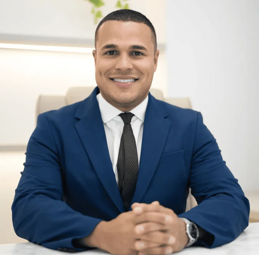 Dr. Marcelo Soares, ortopedista, sorrindo de terno azul-marinho, mãos entrelaçadas sobre mesa de mármore branco, fotografia profissional ultra-realista
