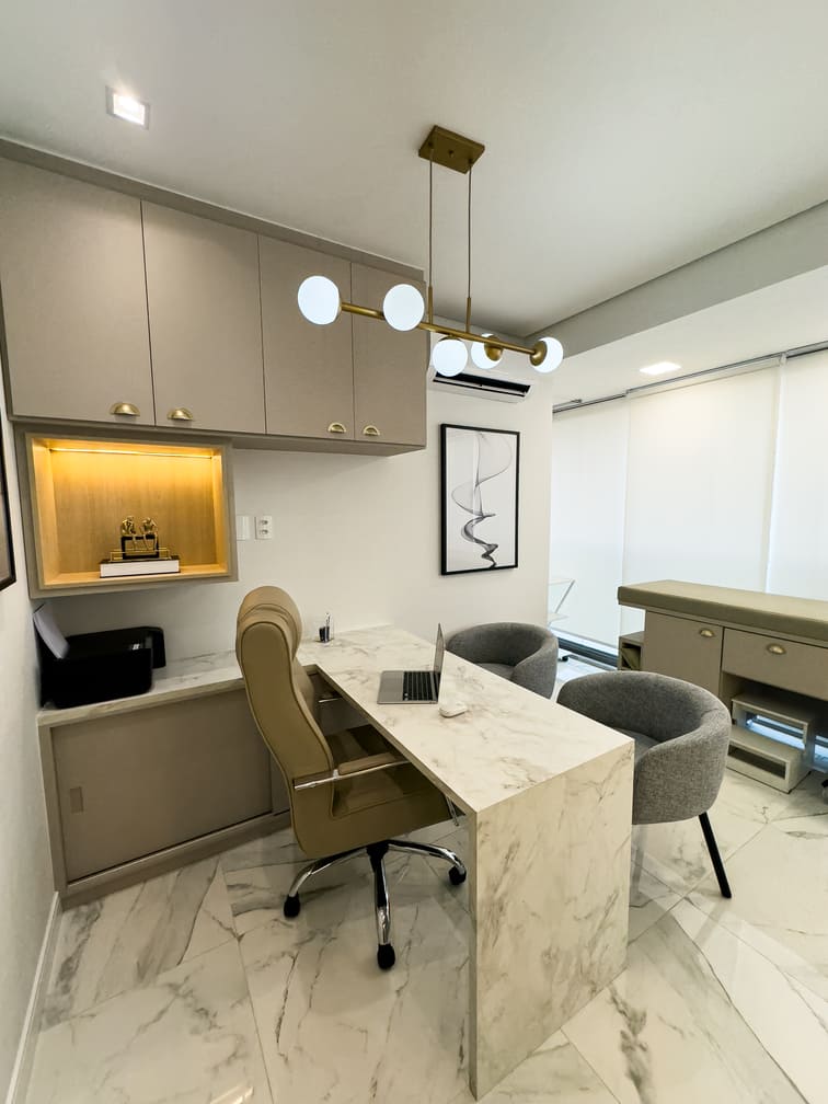 Consultório médico moderno e elegante com mesa de mármore, poltronas cinza, luminária pendente dourada e armários planejados em tom bege, fotografia de interiores ultra-realista